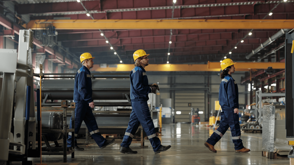 Three warehouse workers wearing yellow hard hats and blue safety uniforms with reflective stripes walking through industrial facility