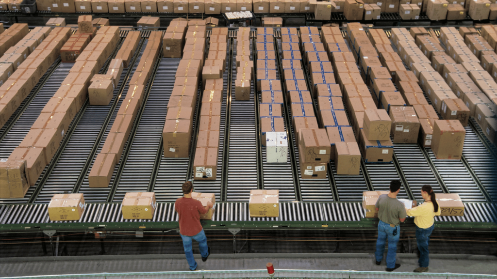 Eye-level shot featuring three people working in a warehouse. The workers are standing in front of moving conveyor belts loaded with numerous boxes, stretching deep into the background. The lighting suggests a bright daytime setting.