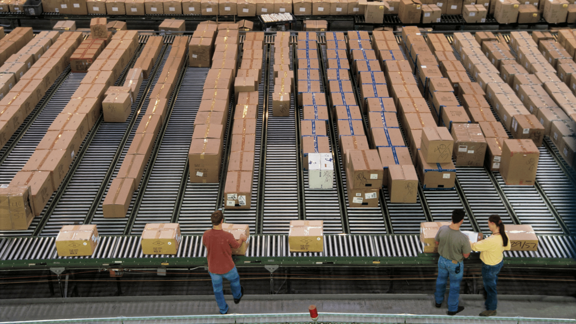 Eye-level shot featuring three people working in a warehouse. The workers are standing in front of moving conveyor belts loaded with numerous boxes, stretching deep into the background. The lighting suggests a bright daytime setting.